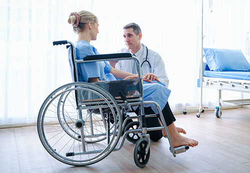 A doctor kneeling and holding the hand of a patient in a wheelchair.